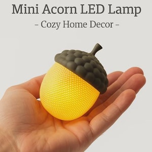 Puede incluir: Mini lámpara LED con forma de bellota, con cuerpo amarillo texturizado y tapa gris oscuro. La lámpara está diseñada para parecerse a una bellota y lleva la etiqueta "Cozy Home Decor". La lámpara se sostiene en una mano.
