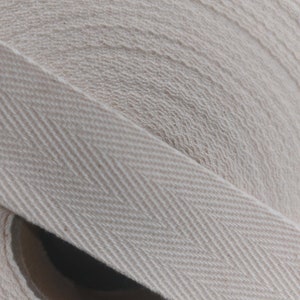 May include: A close-up of a roll of white cotton twill tape with a herringbone pattern. The tape is rolled up and the edges are visible.