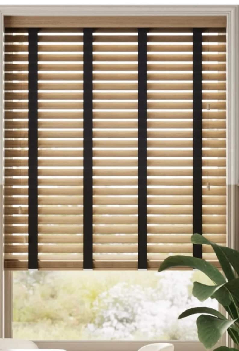May include: Beige wooden blinds with black tape accents. The blinds are closed, but the light shines through the slats.