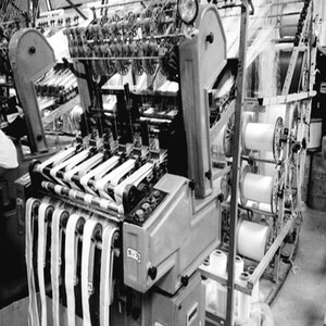 May include: A black and white photograph of a large industrial machine with spools of thread and woven fabric coming out of it.