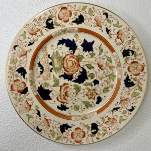 May include: A round ceramic plate featuring a cream-colored base with an intricate floral pattern in shades of burnt orange, dark navy blue, and sage green. The design includes stylized blossoms, scrolling vines, and a central orange flower.