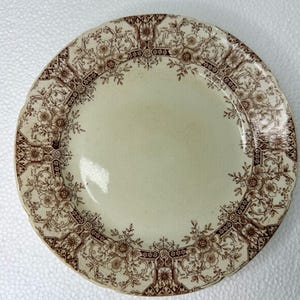 May include: A round ceramic plate with a cream-colored center and a scalloped edge. The rim features a detailed brown transferware pattern consisting of intricate floral motifs, scrolling vines, and geometric architectural elements.