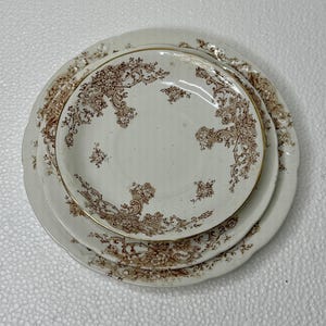 May include: A stack of three round ceramic plates featuring a cream-colored base with intricate brown floral and scrollwork patterns along the scalloped edges. The plates vary in size, with the largest at the bottom and the smallest on top.