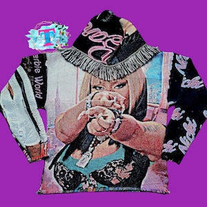 Puede incluir: Una sudadera con capucha de tapicer&iacute;a en blanco y negro con una capucha de flecos rosa y blanca. La sudadera presenta una imagen colorida de una mujer con el texto "Barbie World" y "Nicki" en las mangas.