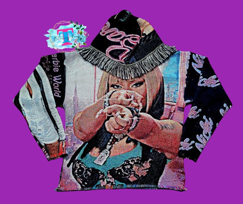 Puede incluir: Una sudadera con capucha de tapicer&iacute;a en blanco y negro con una capucha de flecos rosa y blanca. La sudadera presenta una imagen colorida de una mujer con el texto "Barbie World" y "Nicki" en las mangas.