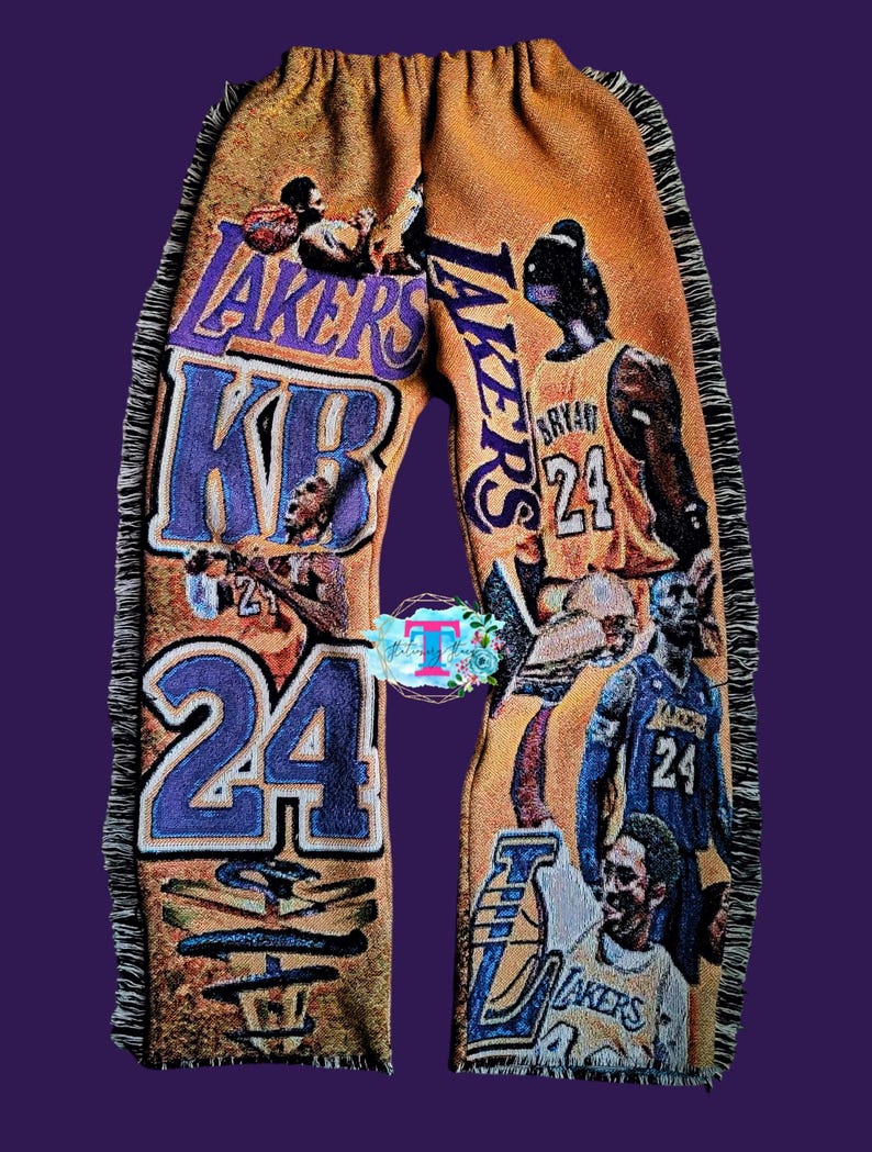 Puede incluir: Un pantal&oacute;n amarillo con ribete de flecos. El pantal&oacute;n est&aacute; cubierto de im&aacute;genes de jugadores de baloncesto que visten camisetas de los Los Angeles Lakers. El texto "LAKERS" y "24" es visible en el pantal&oacute;n.