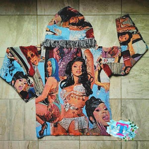 May include: A blue and white hooded sweatshirt with a tapestry design featuring images of women. The sweatshirt has a fringe trim around the hood.