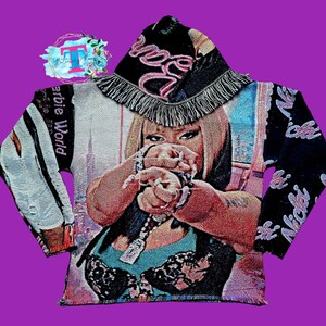 May include: A black and white tapestry hoodie with a pink and white fringe hood. The hoodie features a colorful image of a woman with the text "Barbie World" and "Nicki" on the sleeves.