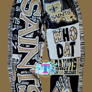Puede incluir: Manta tejida negra y dorada con el logotipo de los New Orleans Saints y las palabras "Who Dat" y "Saints".