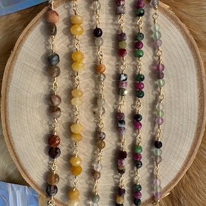 May include: A collection of gemstone necklaces displayed on a wooden slice. Each necklace features a different combination of colorful beads, including yellow, brown, red, green, and purple, strung on delicate gold chains. The necklaces are arranged vertically, showcasing their unique designs.