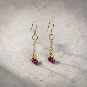 May include: A pair of gold-colored dangle earrings. Each earring features a hook, a small circle, a delicate chain, and two small, round, deep red beads at the bottom. The earrings are displayed against a light gray background.