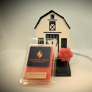 Puede incluir: Un calentador de cera en forma de granero blanco con detalles negros y una cera perfumada roja. La cera perfumada está etiquetada como "Apple Pecan Sage" y está en un recipiente de plástico transparente. El calentador tiene una base negra y una flor roja decorativa.