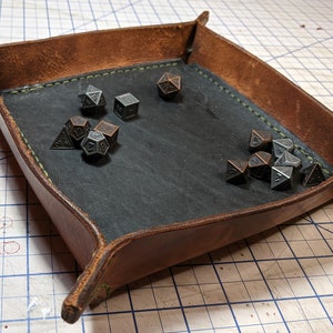 Könnte beinhalten: Ein braunes Leder-Würfeltablett mit schwarzem Filz-Innenraum. Das Tablett hat einen grünen, genähten Rand und ist mit Metallwürfeln gefüllt.