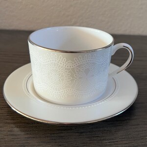 May include: White porcelain teacup and saucer set with a delicate white lace pattern and a silver rim.