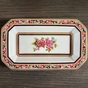 Puede incluir: Una bandeja rectangular de cerámica blanca con un borde dorado y un diseño floral negro y rojo. La bandeja tiene un borde festoneado y es perfecta para servir aperitivos o joyas.