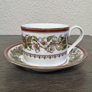 May include: White teacup and saucer set with a floral and holly design. The cup has a red band with gold trim, and the saucer mirrors the cup's design. The set is likely porcelain and suitable for serving tea or coffee.