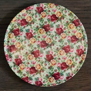 May include: A round, white ceramic coaster with a red and yellow floral pattern. The coaster features a repeating design of roses and leaves.