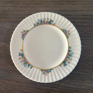 May include: A white ceramic plate with a scalloped edge and a floral design in shades of blue, green, yellow, and purple. The plate has a gold rim.