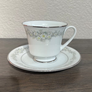 May include: A white teacup and saucer set with a delicate floral pattern of white daisies and green leaves. The cup has a gold rim and the saucer has a gold trim.
