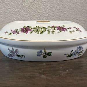 May include: White oval ceramic baking dish with a floral design. The dish has a lid with a gold trim and a floral design. The flowers are purple, pink, and blue.