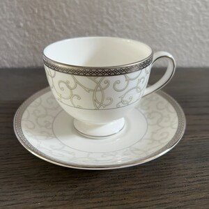 May include: A white teacup and saucer set with a silver and gold floral pattern. The cup and saucer have a silver rim.