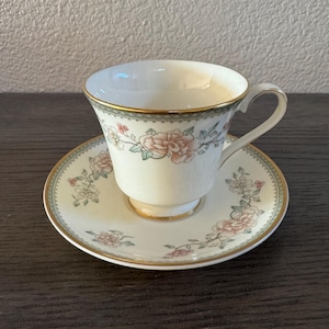 May include: Cream-colored teacup and saucer set with a floral design. The cup and saucer have a gold trim and a delicate pattern of pink and green flowers. The teacup has a curved handle.