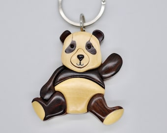 Puede incluir: Un llavero de madera con forma de panda con la cara en blanco y negro y el pelo marrón. El panda está sentado y saludando con su pata derecha.