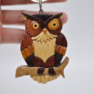 May include: Wooden owl keychain with intricate details, featuring a brown, orange, and yellow color scheme. The owl is perched on a branch and has large, round eyes.
