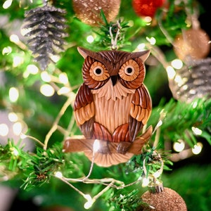 May include: A wooden owl ornament with brown, orange, and white details, hanging on a green Christmas tree with gold lights and other ornaments.