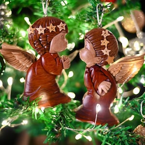 May include: Two wooden angel ornaments with wings, brown and red paint, and gold stars on their heads, hanging on a Christmas tree with green branches and white lights.