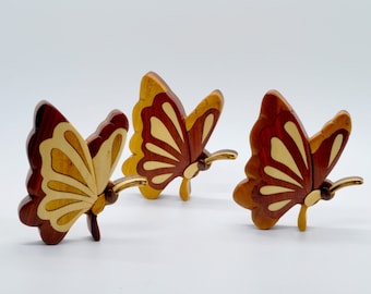 Puede incluir: Tres adornos de madera en forma de mariposa con detalles intrincados. Las mariposas están hechas de madera de diferentes tonos de marrón y amarillo.