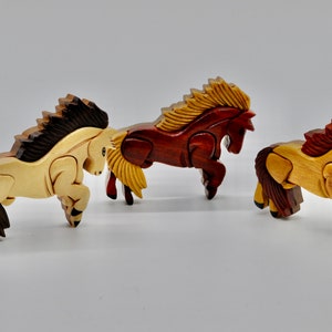 K&ouml;nnte beinhalten: Drei h&ouml;lzerne Pferdefiguren, jede aus einem anderen Holz geschnitzt. Die Pferde sind in einer springenden Pose, mit ihren M&auml;hnen und Schw&auml;nzen, die hinter ihnen wehen.