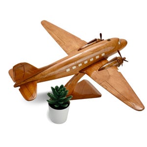 K&ouml;nnte beinhalten: Ein h&ouml;lzernes Flugzeugmodell mit hellbrauner Oberfl&auml;che. Das Flugzeug hat eine kleine gr&uuml;ne Pflanze in einem wei&szlig;en Topf davor.