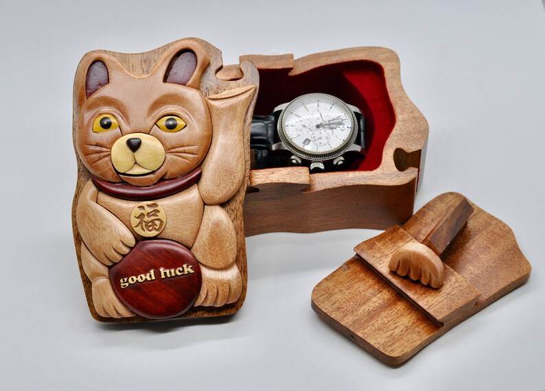 以下が含まれることがあります： 赤い内装と時計が入った招き猫の形をした木製の箱。箱にはスライド式の蓋と側面に小さな木製の足があります。箱には「幸運」という言葉と中国の文字が刻まれています。