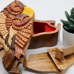 Op de afbeelding: Een houten uil puzzeldoos met een rood fluwelen interieur. De doos is ingewikkeld gesneden met twee uilen die op een tak zitten. De deksel van de doos is open, waardoor het interieur zichtbaar is.