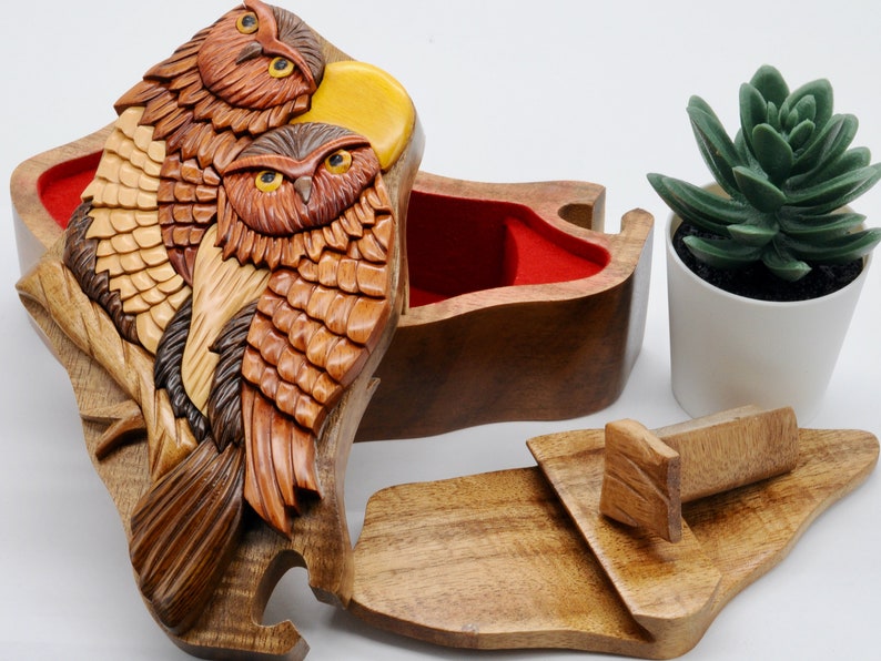 Op de afbeelding: Een houten uil puzzeldoos met een rood fluwelen interieur. De doos is ingewikkeld gesneden met twee uilen die op een tak zitten. De deksel van de doos is open, waardoor het interieur zichtbaar is.