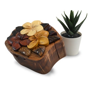 May include: A wooden puzzle box featuring a carved design of tropical flowers. The box is crafted from dark wood and has a natural finish. The flowers are carved in various shades of brown, yellow, and red.