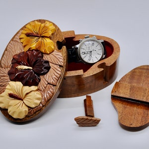 May include: A wooden puzzle box with a carved hibiscus flower design. The box is open and a watch is inside. The box has a separate lid and a small wooden piece that fits into the box.