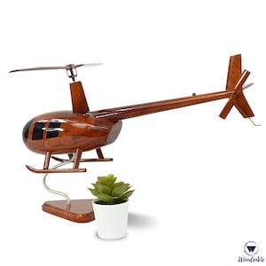 May include: A handcrafted wooden helicopter model with a rich, dark brown finish. The model features detailed rotor blades, a tail rotor, and a sleek fuselage. The helicopter is displayed on a silver stand, with a small potted succulent nearby.