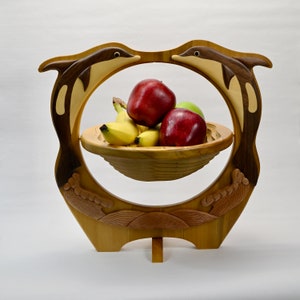 Op de afbeelding: Een houten fruitschaal met een dolfijnontwerp. De schaal wordt ondersteund door een standaard en heeft een cirkelvormige opening met twee dolfijnen die tegenover elkaar staan. De schaal bevat rode appels, groene appels en bananen. De dolfijnen zijn bruin en beige.