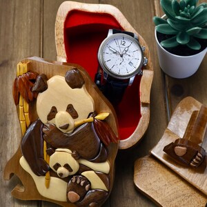 Puede incluir: Una caja de rompecabezas de madera con un panda tallado y su cachorro. La caja est&aacute; abierta y un reloj plateado con una correa de cuero negro est&aacute; en el interior. La caja est&aacute; forrada con terciopelo rojo.
