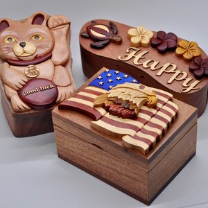 以下が含まれることがあります： 複雑な彫刻が施された木製の箱パズルが3つあります。最初の箱は、前面に「幸運」と書かれた茶色の猫が描かれています。2番目の箱は波の形をしており、前面に「Happy」という文字が刻まれています。3番目の箱は、上にワシが乗ったアメリカ国旗が描かれています。