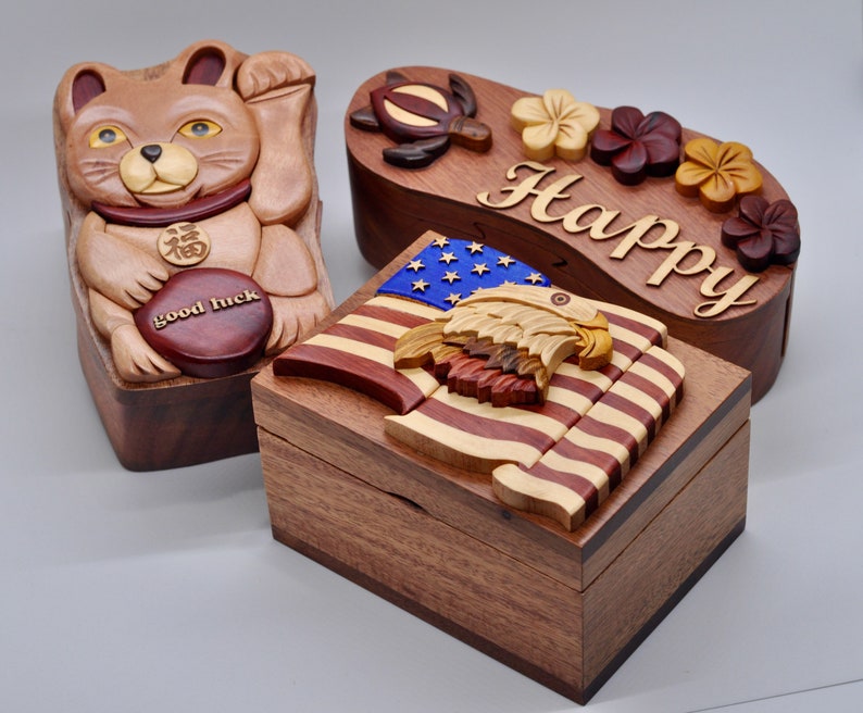 以下が含まれることがあります： 複雑な彫刻が施された木製の箱パズルが3つあります。最初の箱は、前面に「幸運」と書かれた茶色の猫が描かれています。2番目の箱は波の形をしており、前面に「Happy」という文字が刻まれています。3番目の箱は、上にワシが乗ったアメリカ国旗が描かれています。