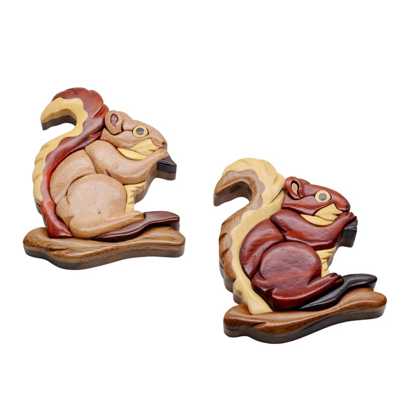 May include: Two wooden squirrel figurines. One is light brown with a reddish-brown tail, the other is dark brown with a lighter tail. Both are holding a dark object and sitting on a wooden base. The squirrels are carved with detailed features.