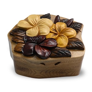May include: A wooden puzzle box featuring a carved design of three plumeria flowers in shades of brown, yellow, and red. The box is closed and has a smooth, polished finish.