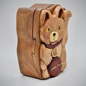 以下が含まれることがあります： 茶色の猫の形をした木製の箱パズル。猫は黄色い目を持ち、胸には「幸運」の中国語の文字が書かれた金貨があります。箱は赤い木製の土台になっており、「幸運」の文字が刻まれています。