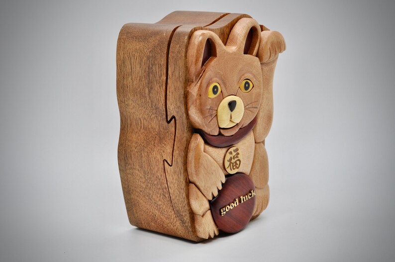 以下が含まれることがあります： 茶色の猫の形をした木製の箱パズル。猫は黄色い目を持ち、胸には「幸運」の中国語の文字が書かれた金貨があります。箱は赤い木製の土台になっており、「幸運」の文字が刻まれています。