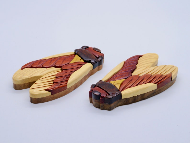 Puede incluir: Dos cajas de rompecabezas de madera con forma de cigarra con detalles intrincados. Las cajas est&aacute;n hechas de diferentes tipos de madera, incluyendo arce, nogal y cerezo. Las alas est&aacute;n talladas con detalles realistas y los cuerpos est&aacute;n pintados con una mancha marr&oacute;n oscura.