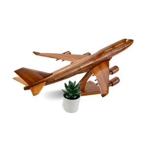 May include: A wooden model airplane with four engines, wings, and a tail, displayed on a wooden stand. The airplane is a warm brown color. A small green succulent in a white pot is in front of the airplane.