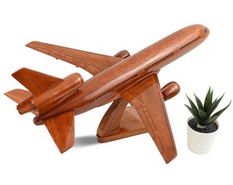 Könnte beinhalten: Ein poliertes Flugzeugmodell aus Holz mit Tri-Jet-Design und glänzender Oberfläche, montiert auf einem passenden Holzständer. Das Flugzeug zeigt eingravierte Fenster- und Türdetails entlang des Rumpfes und der Flügel. Eine kleine grüne Sukkulente in einem weißen Topf.
